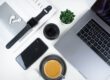 The image depicts a modern, minimalist workspace setup, featuring a laptop, smartphone, wireless earbuds, a notepad, a black stylus or pen, an analog wristwatch, a cup of coffee on a saucer, an open magazine or catalog, and a camera lens.