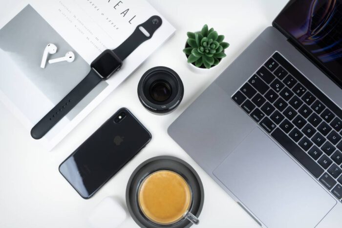 The image depicts a modern, minimalist workspace setup, featuring a laptop, smartphone, wireless earbuds, a notepad, a black stylus or pen, an analog wristwatch, a cup of coffee on a saucer, an open magazine or catalog, and a camera lens.