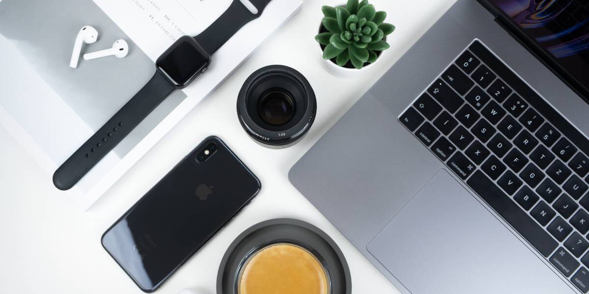 img020 The image depicts a modern, minimalist workspace setup, featuring a laptop, smartphone, wireless earbuds, a notepad, a black stylus or pen, an analog wristwatch, a cup of coffee on a saucer, an open magazine or catalog, and a camera lens.